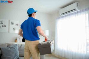 A man in a blue shirt carrying a box in a living room, illustrating the importance of a comfortable home environment with proper garage AC repair.