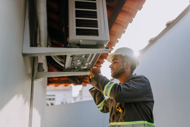A man wearing a hard hat and safety vest is servicing a wall AC unit.