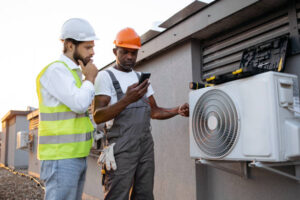 Two men wearing hard hats and safety vests examining a cell phone, discussing maintenance of AC systems.